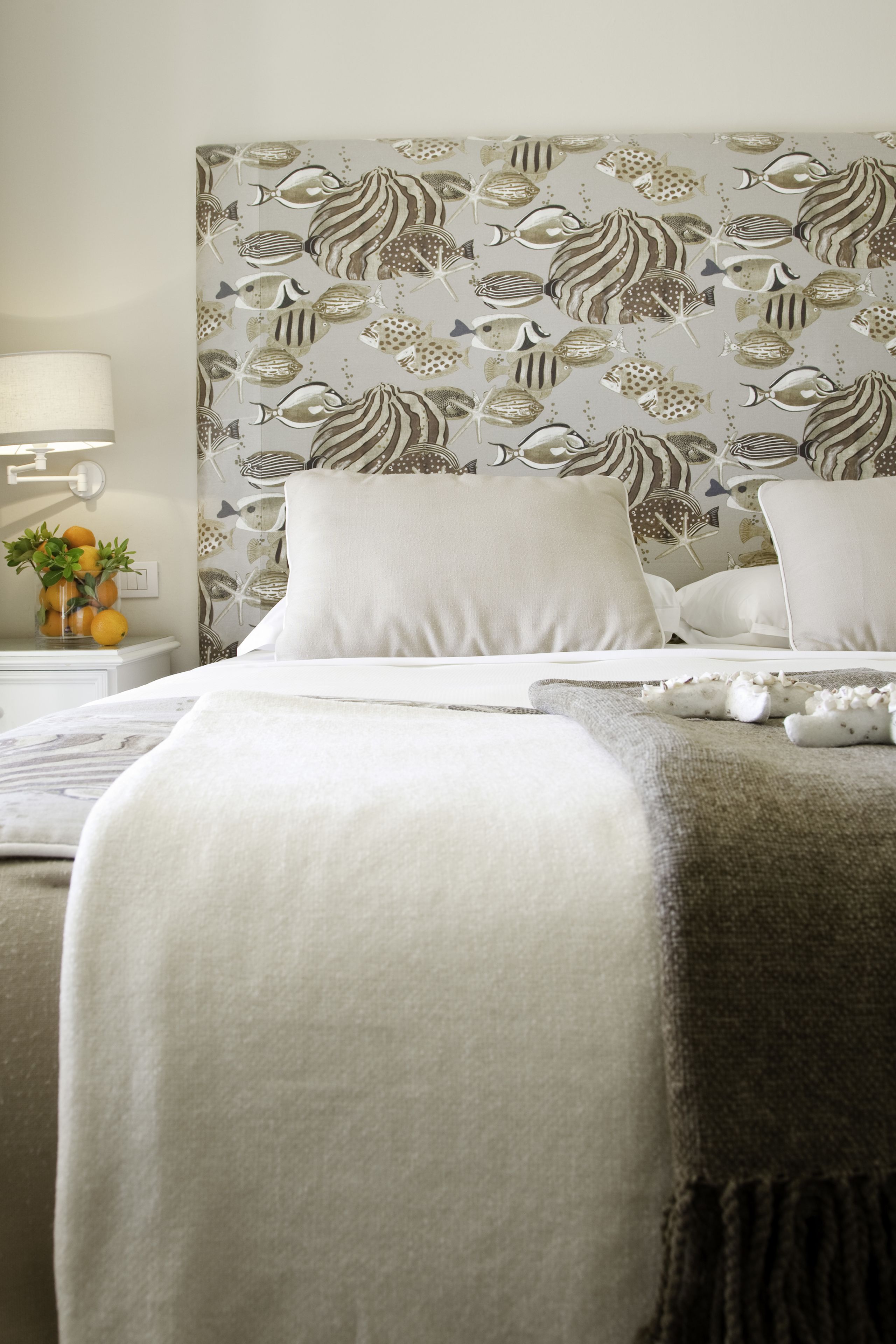 Un letto rifatto con lenzuola bianche, una testiera decorata e un comodino con una lampada e un vaso di fiori.