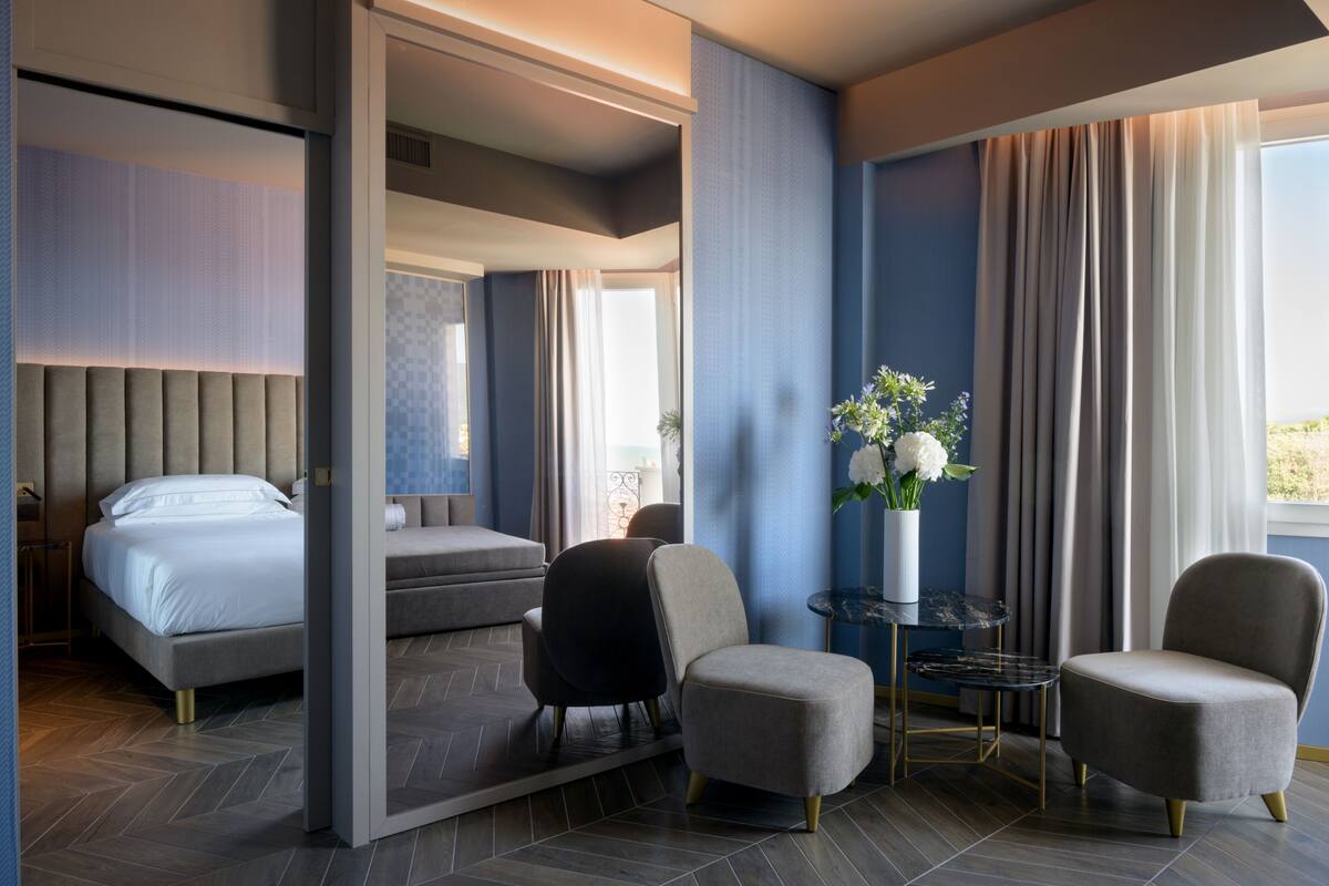 A modern hotel room with a large bed, two armchairs, a small table, and a mirror.