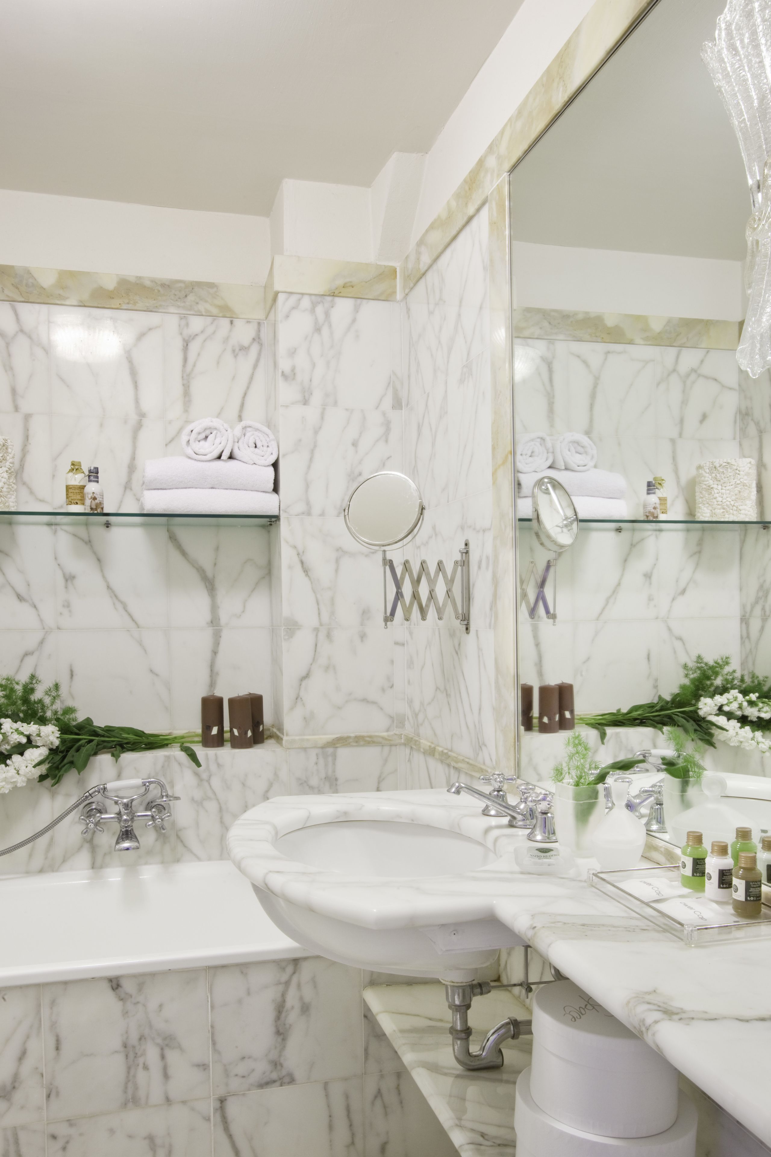 Un baño con paredes de mármol, dos lavamanos, un espejo grande y estantes con artículos de aseo.