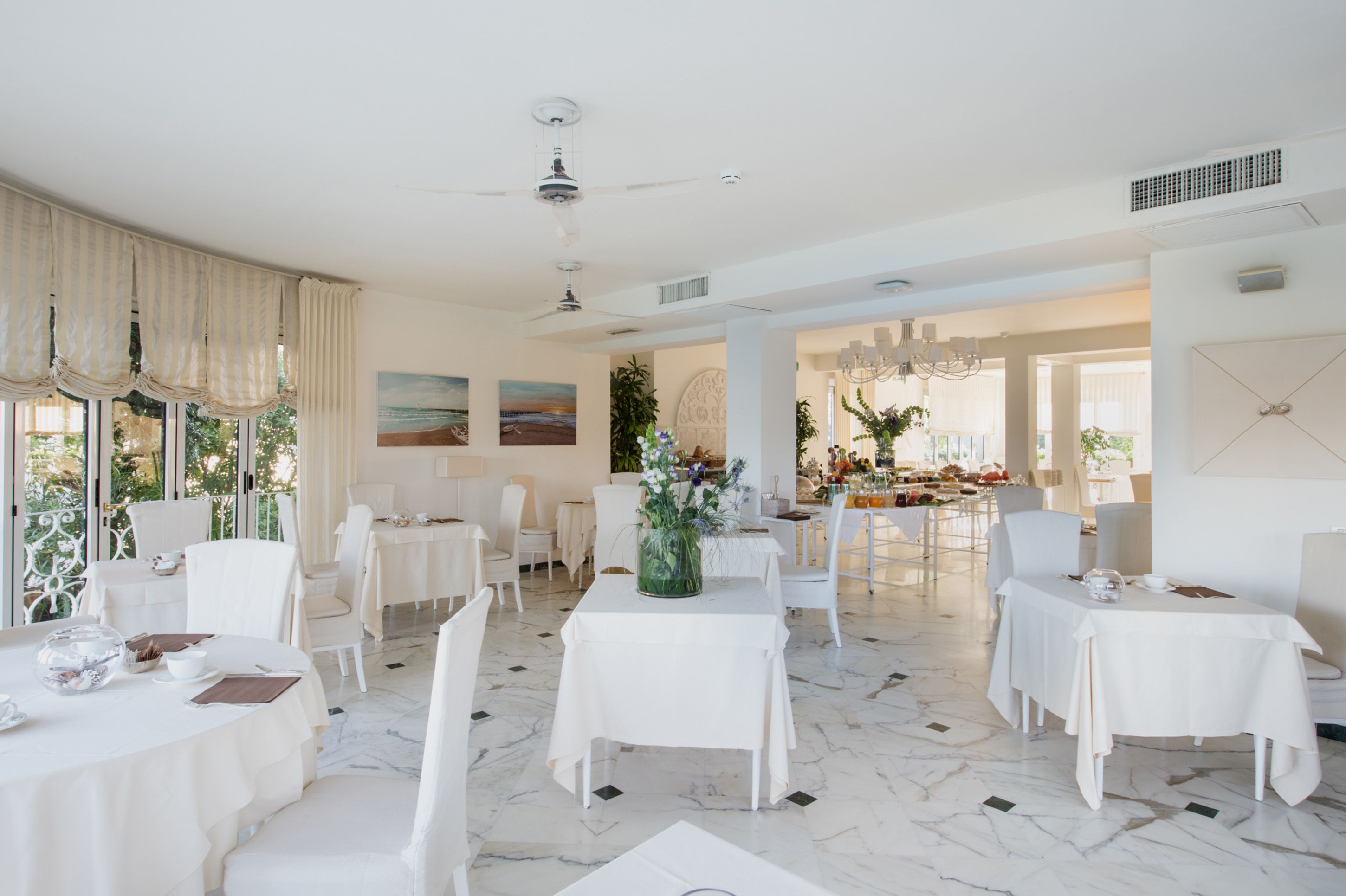Un comedor amplio con mesas y sillas blancas, un ventilador de techo y vistas a la terraza exterior.