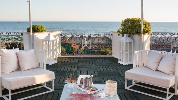 A terrace with two white cushioned chairs, a table with a basket of food and drinks, and a view of the sea and buildings. - Hotel Il Negresco (Forte dei Marmi)