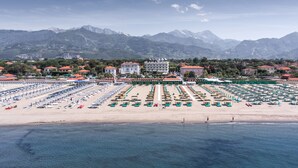 A beach with rows of sunbeds and umbrellas, a building with a red roof, and mountains in the background. - Hotel Il Negresco (Forte dei Marmi)