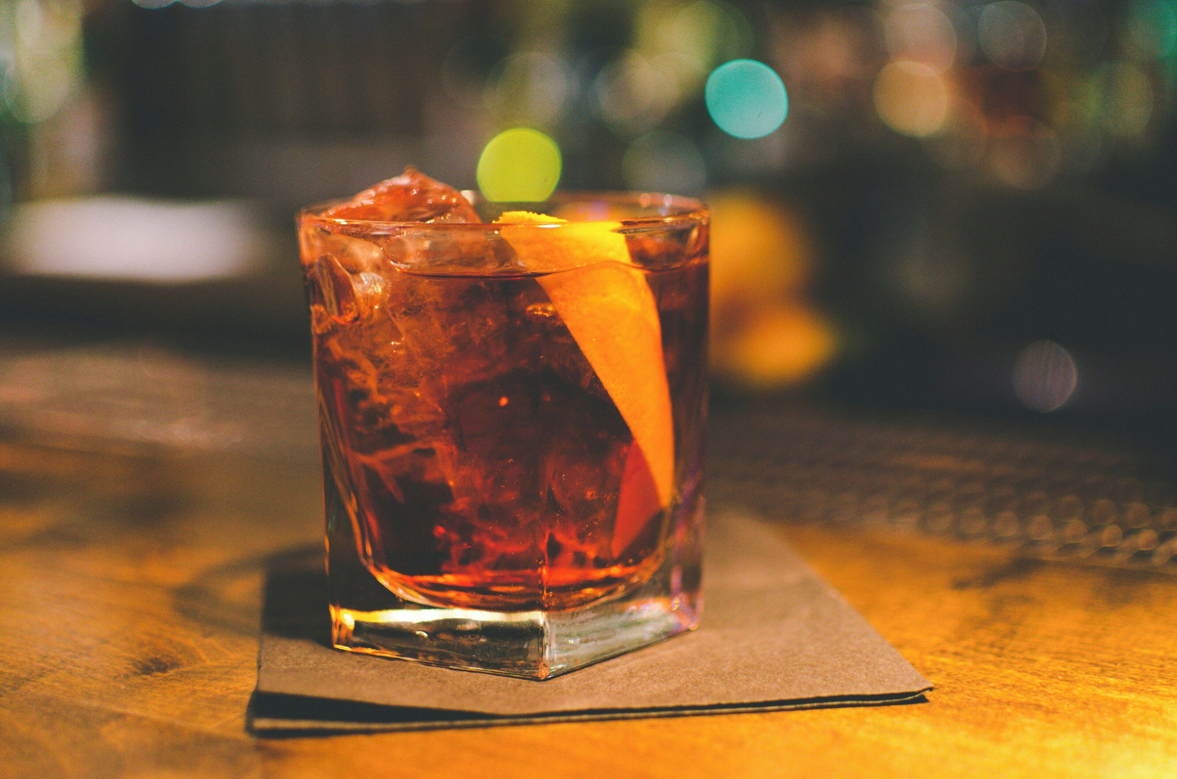 Un verre d’une boisson ambrée, contenant des glaçons et une tranche de zeste d’orange, posé sur une table en bois avec une serviette en dessous.