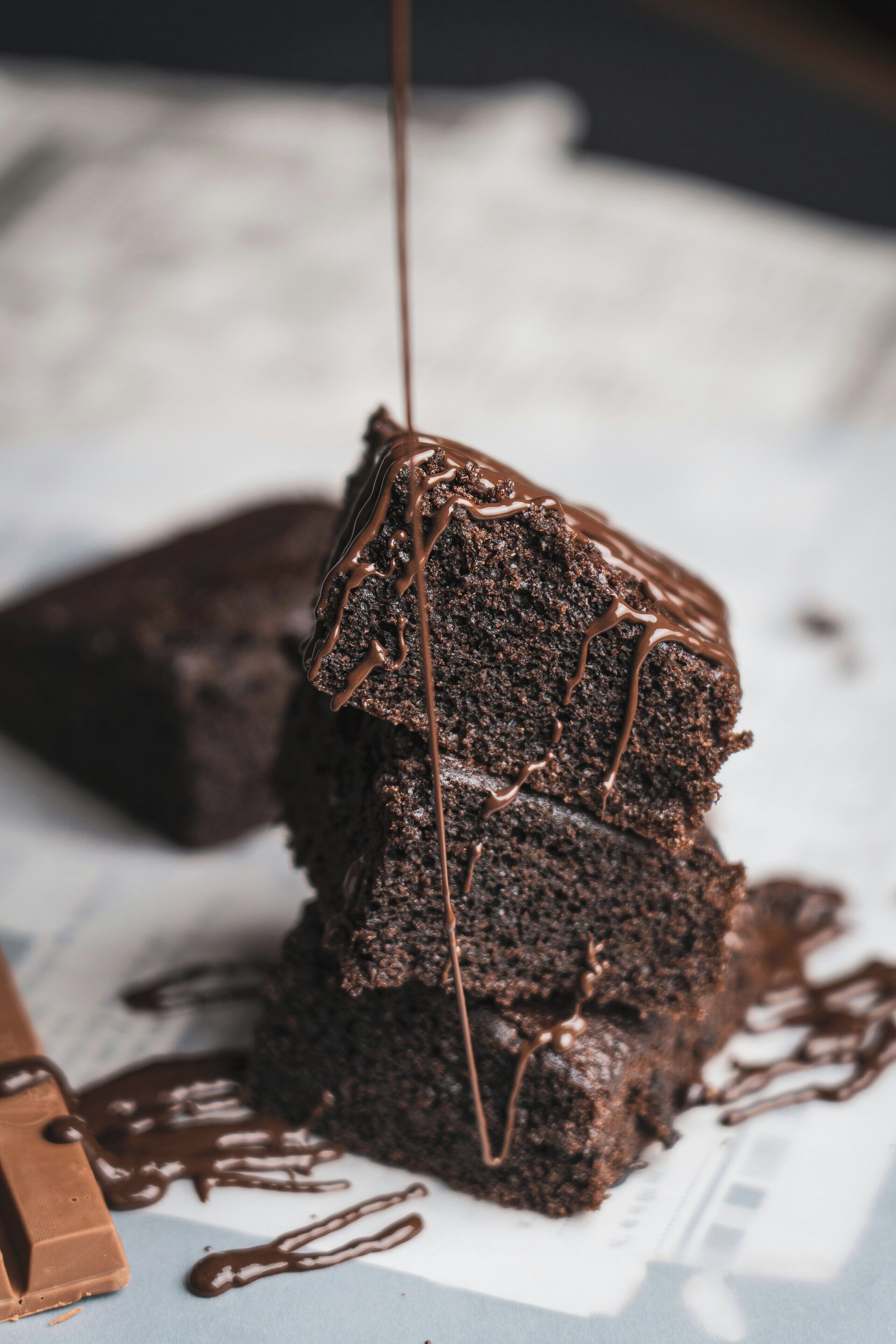 A stack of chocolate brownies with chocolate sauce dripping down, placed on a white surface with a knife beside them.