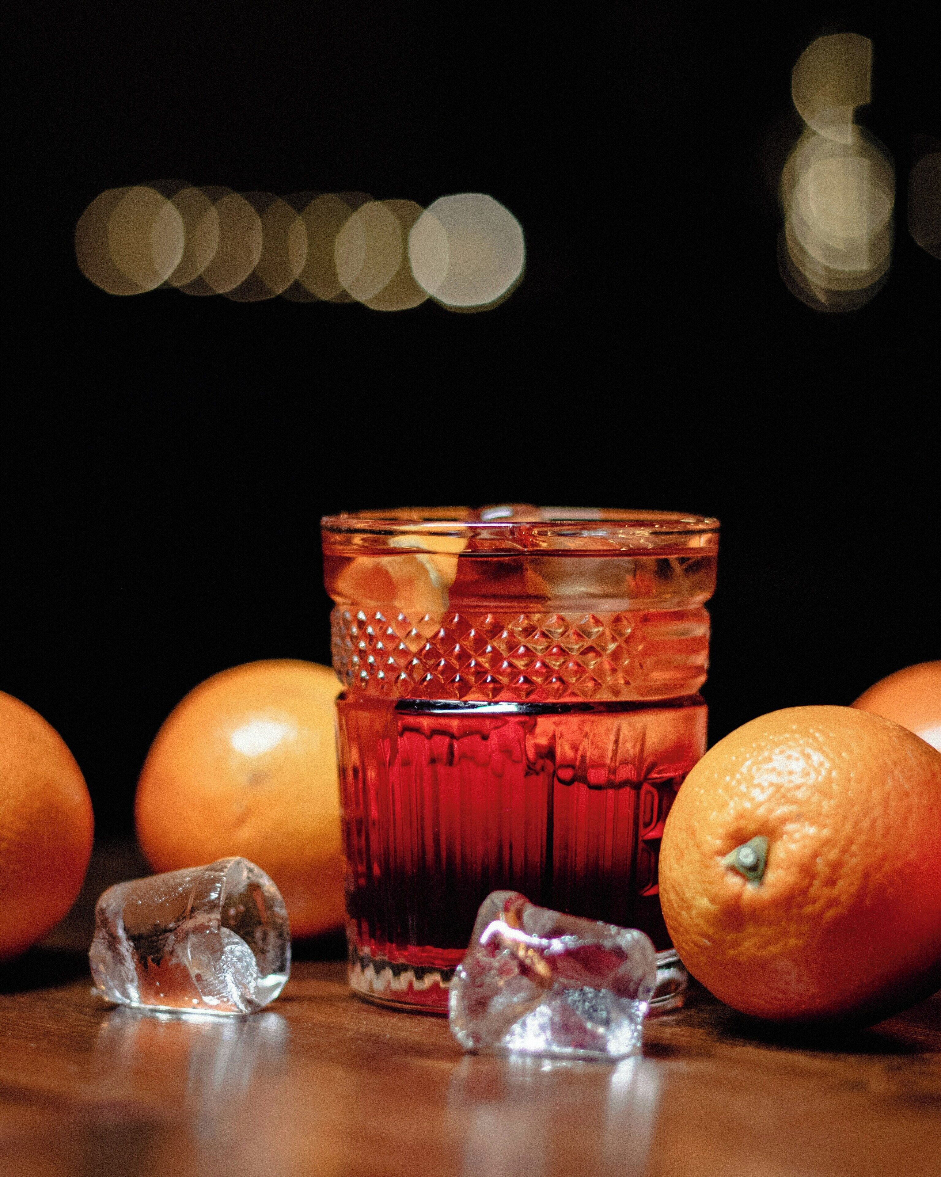 Un verre contenant des glaçons et une tranche d’orange, entouré d’oranges entières sur une surface en bois.