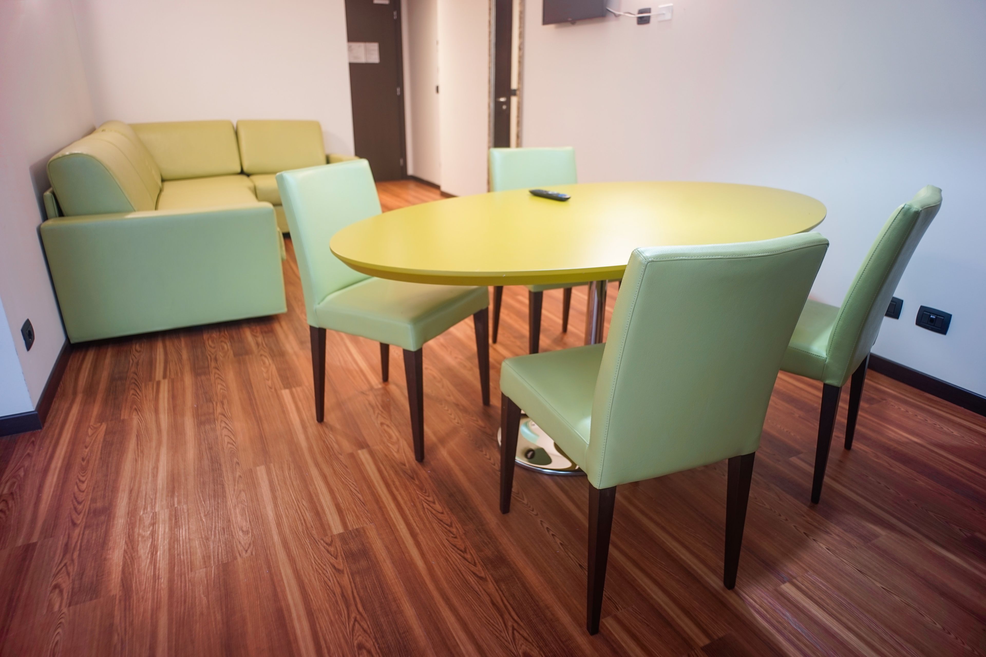 A room with a round yellow table and four light green chairs, a beige sofa, and wooden flooring.