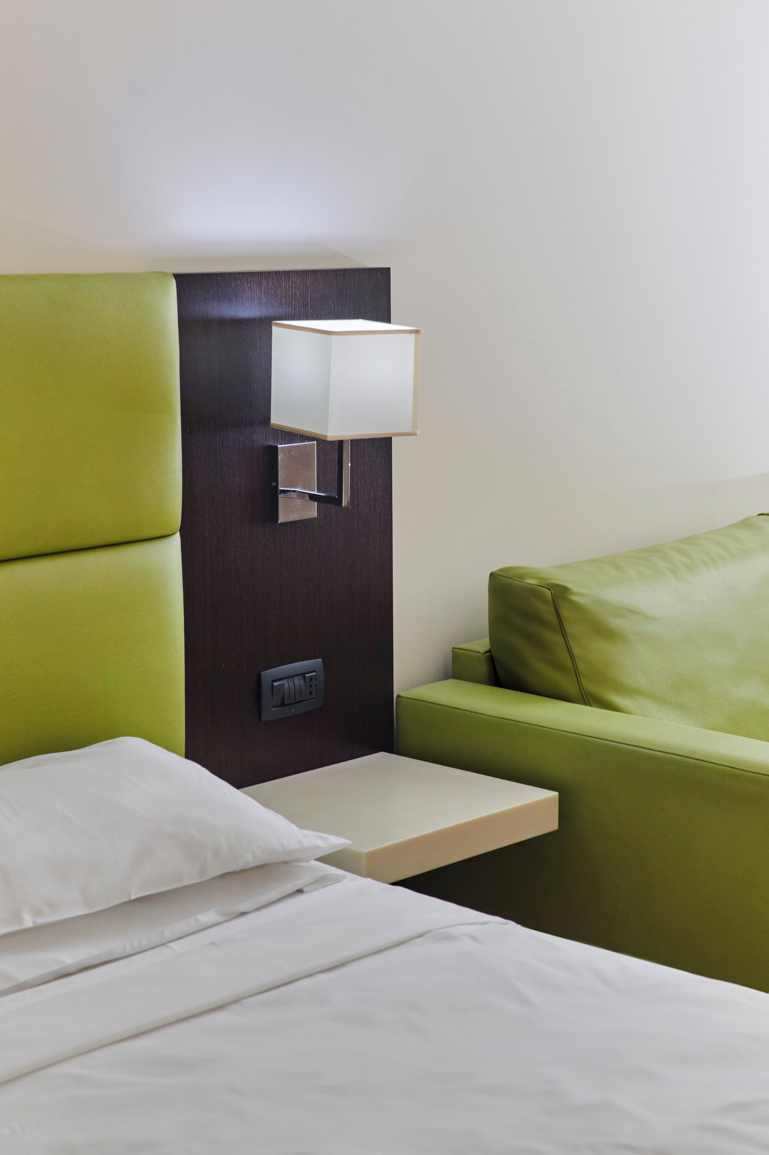Une chambre d’hôtel avec un lit, une tête de lit recouverte d’un tissu vert, une table de chevet avec une lampe et une prise de courant.