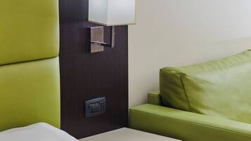 Une chambre d’hôtel avec un lit, une tête de lit recouverte d’un tissu vert, une table de chevet avec une lampe et une prise de courant.