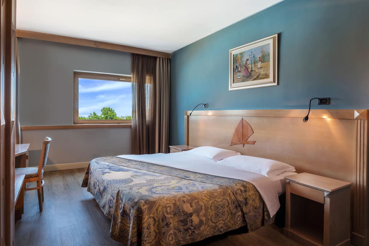 A hotel room with a large bed, a desk, a chair, a window with curtains, a painting on the wall, and a nightstand.
