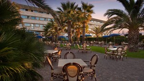 Un patio al aire libre con mesas y sillas, rodeado de palmeras y un edificio al fondo.