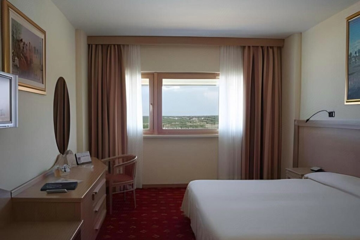A hotel room with a bed, a desk, a chair, a window with curtains, and a painting on the wall.