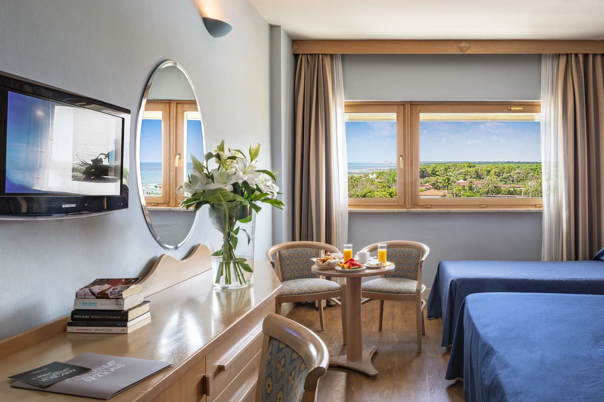A hotel room with two beds, a TV, a dining table with chairs, a vase with flowers, and a view of the sea and greenery.