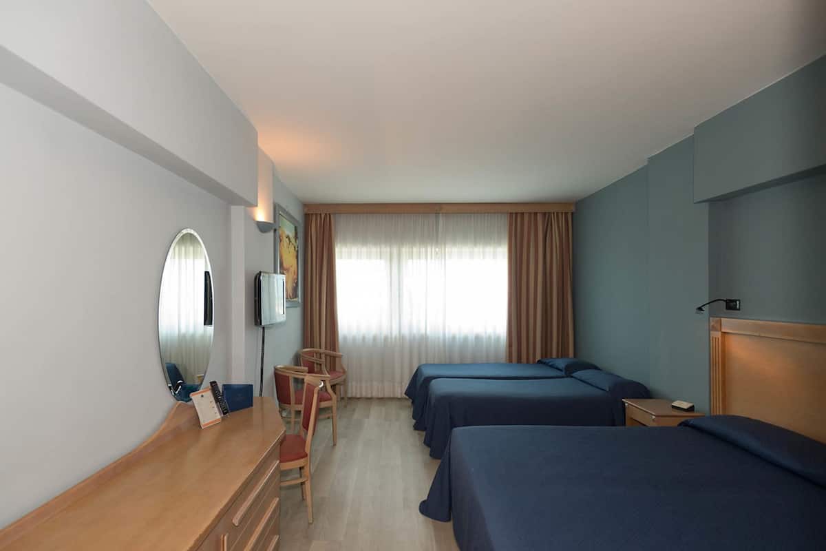 A hotel room with a wooden desk, a chair, a bed, a TV, and a window with curtains.