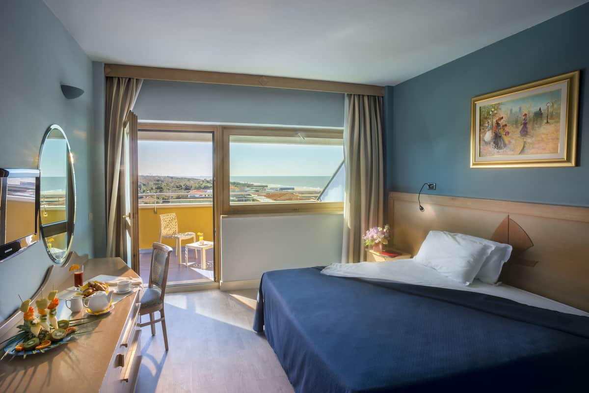 A hotel room with a bed, a dining table with breakfast, a chair, a large window with a view, and a painting on the wall.
