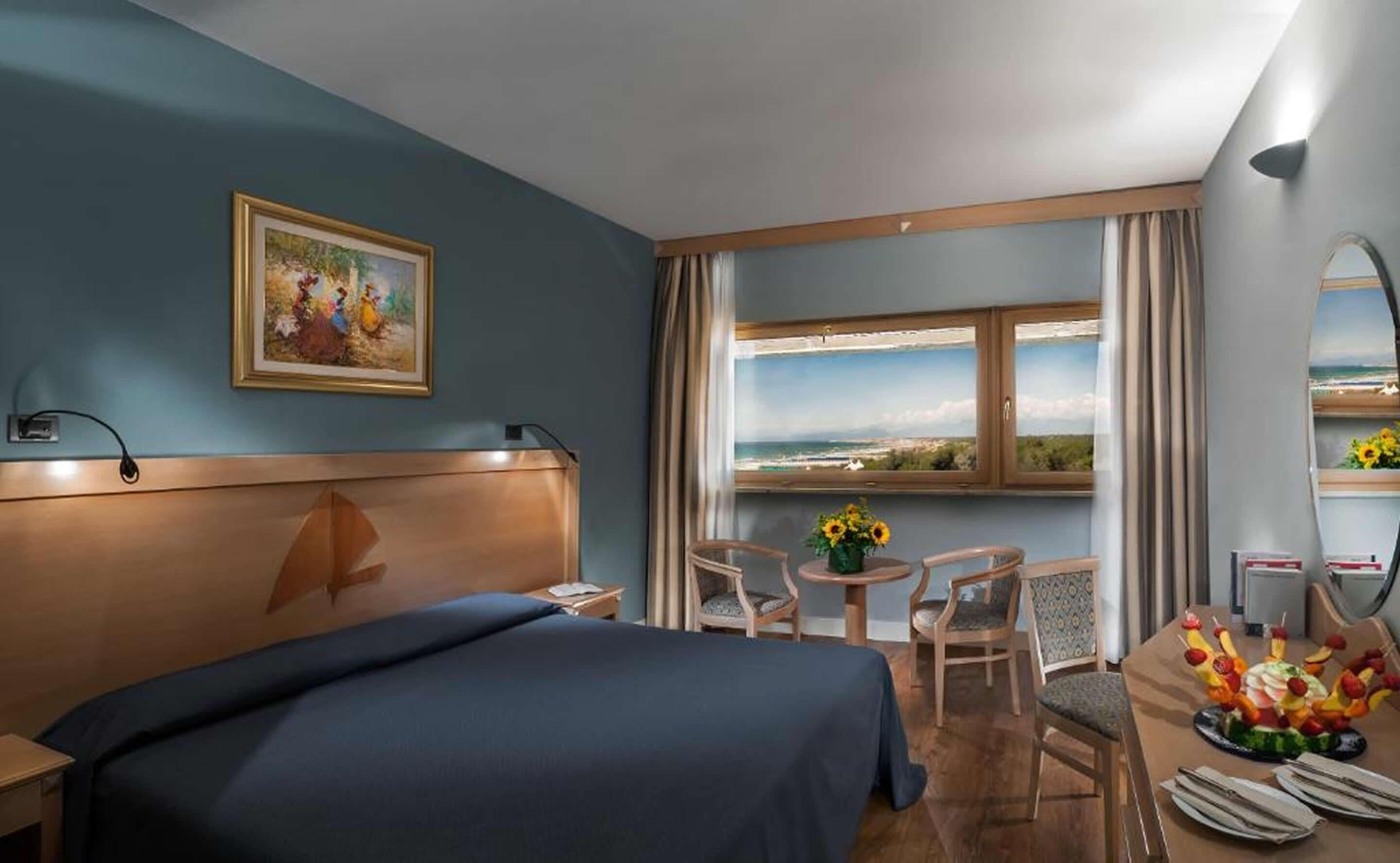 Une chambre d’hôtel avec un lit, un bureau agrémenté d’un vase de fleurs, une chaise, une vue sur l’océan et un tableau au mur.