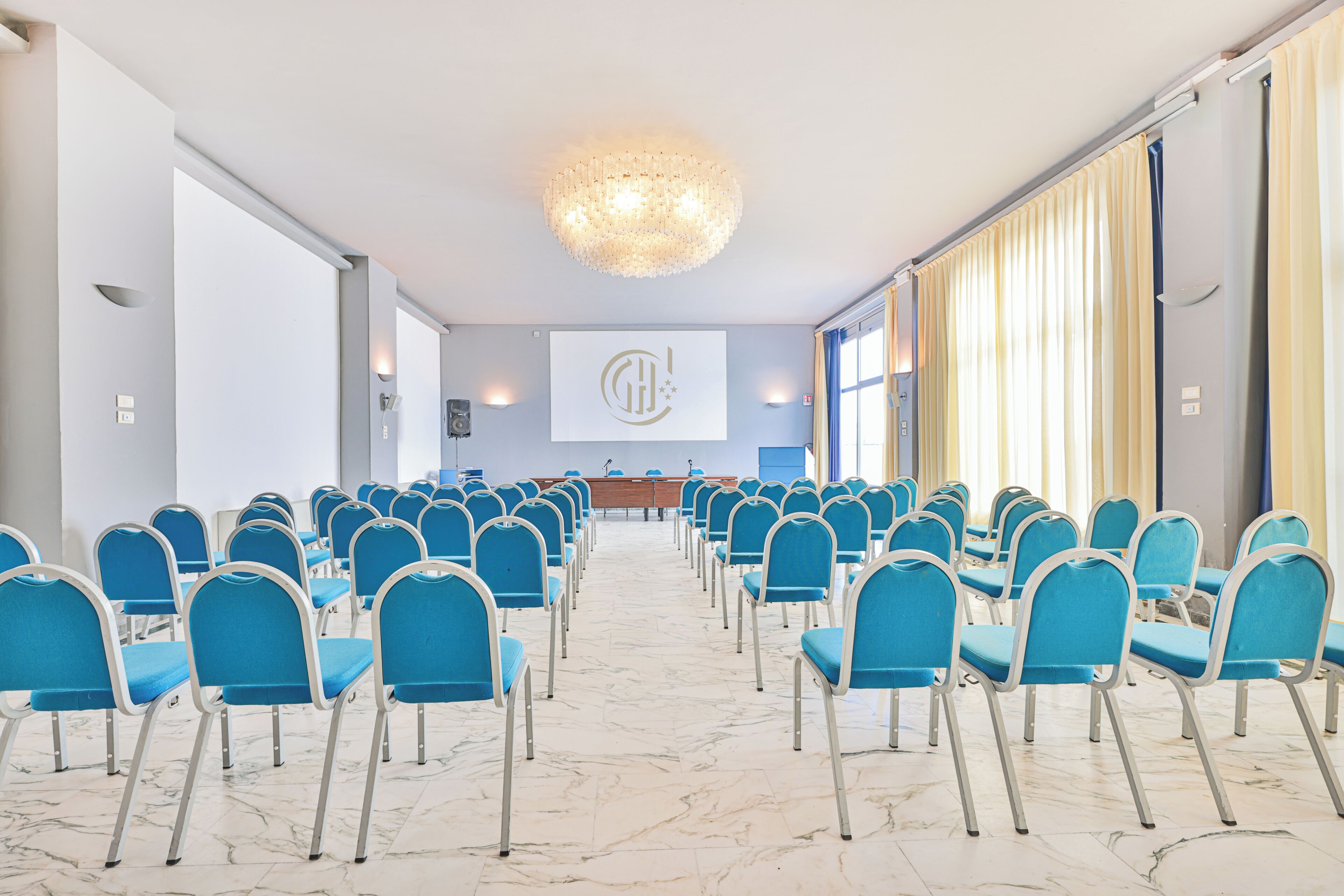 A conference room with rows of blue chairs, a marble floor, a large screen with a logo, and a chandelier.
