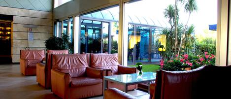 Un vestíbulo amplio con sofás de cuero, una mesa de centro de vidrio y grandes ventanas con vistas a un jardín y palmeras.
