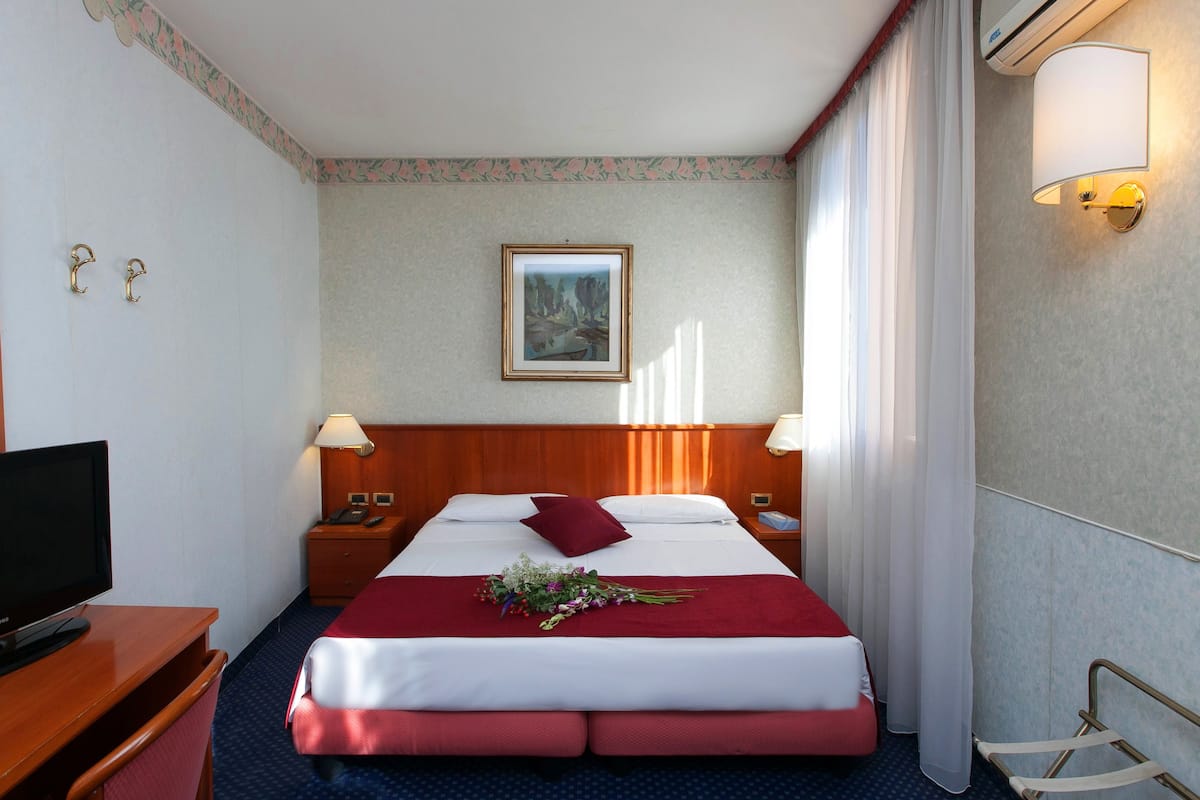 A hotel room with a bed, a desk with a TV, a nightstand, a wall-mounted lamp, and a framed picture on the wall.
