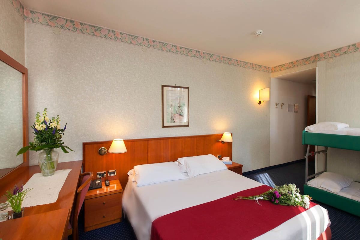 A hotel room with a bed, bedside tables, a mirror, a picture on the wall, and a bunk bed in the corner.