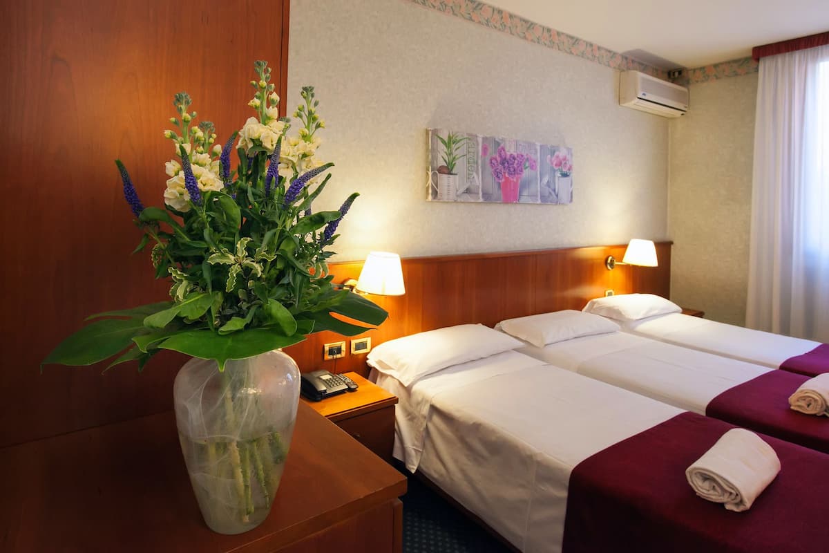 A hotel room with two beds, a wooden headboard, a nightstand with a phone, a vase with flowers, and a painting on the wall.