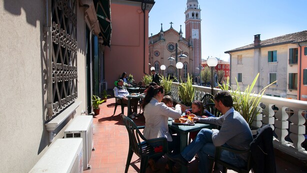 Un café en plein air avec des tables et des chaises, un bâtiment doté d’une tour horloge et un ciel dégagé.