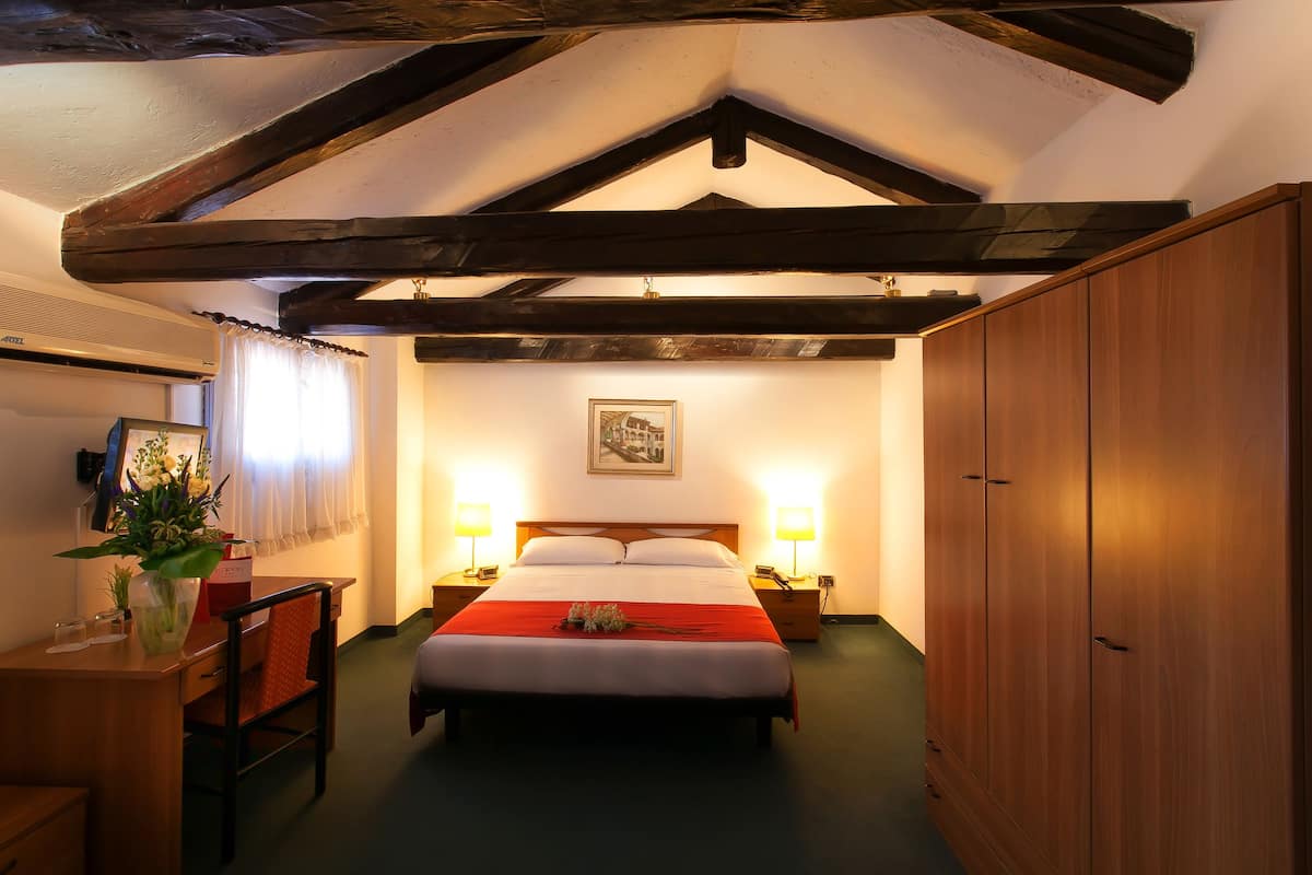 A hotel room with a wooden ceiling, a bed with a red blanket, two bedside lamps, a desk with a chair, and a large wardrobe.