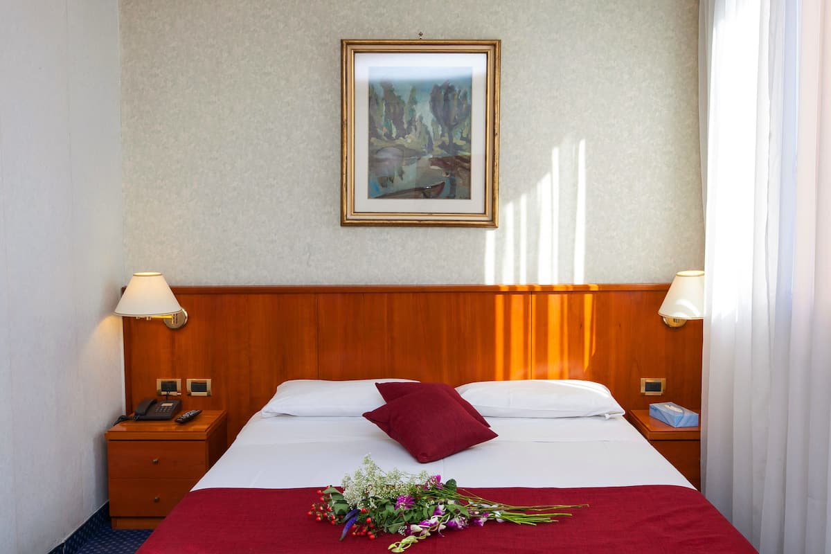 A hotel room with a wooden headboard, a framed artwork on the wall, a red throw blanket, a bouquet of flowers, and two bedside lamps.