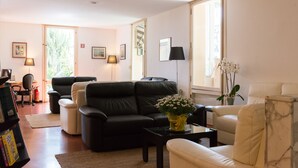 Un salon avec un canapé en cuir noir, un fauteuil blanc, une table basse et un vase rempli de fleurs. On y trouve également une bibliothèque avec des livres et une lampe sur une table d’appoint.