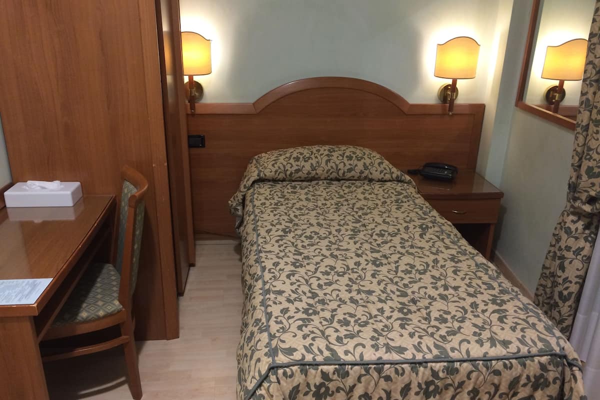 Ein Hotelzimmer mit einem hölzernen Kopfteil, einem Einzelbett mit Blumenmuster auf der Bettwäsche, einem Nachttisch mit Lampe, einem Sessel und einem Schreibtisch mit Taschentuchbox.