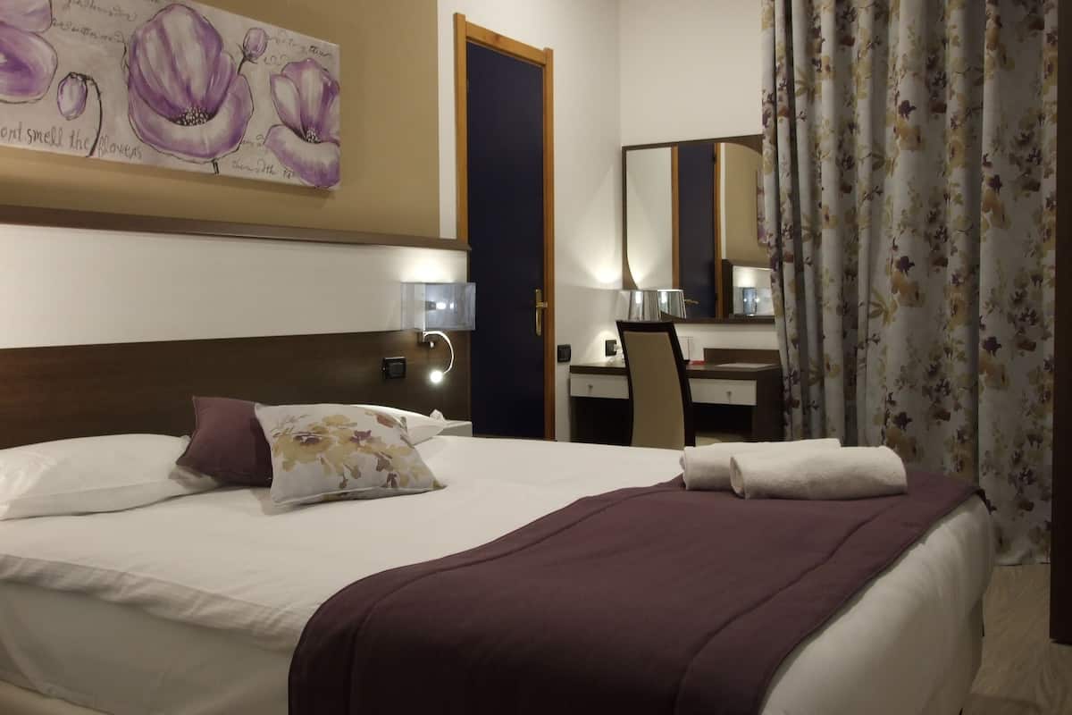 Ein Hotelzimmer mit einem großen Bett, einem Schreibtisch, einem Sessel, einem Spiegel und einem Blumenbild an der Wand.