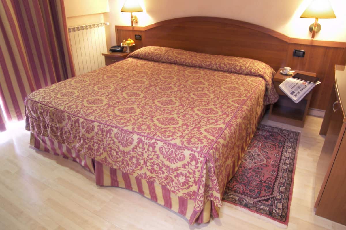 Ein Hotelzimmer mit einem großen Bett, zwei Nachttischlampen, einem Nachttisch, einem kleinen Tisch mit einer Zeitung und einem Teppich.