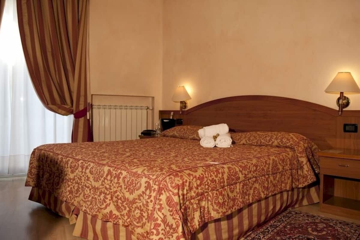 Ein Hotelzimmer mit einem großen Bett, zwei Nachttischlampen, einem Holz-Kopfteil, einem Nachttisch und einem Fenster mit Vorhängen.
