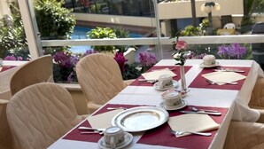 Une table à manger dressée avec des nappes blanches, des chaises de couleur beige, et une vue sur une piscine et la verdure, visible à travers une fenêtre en verre.