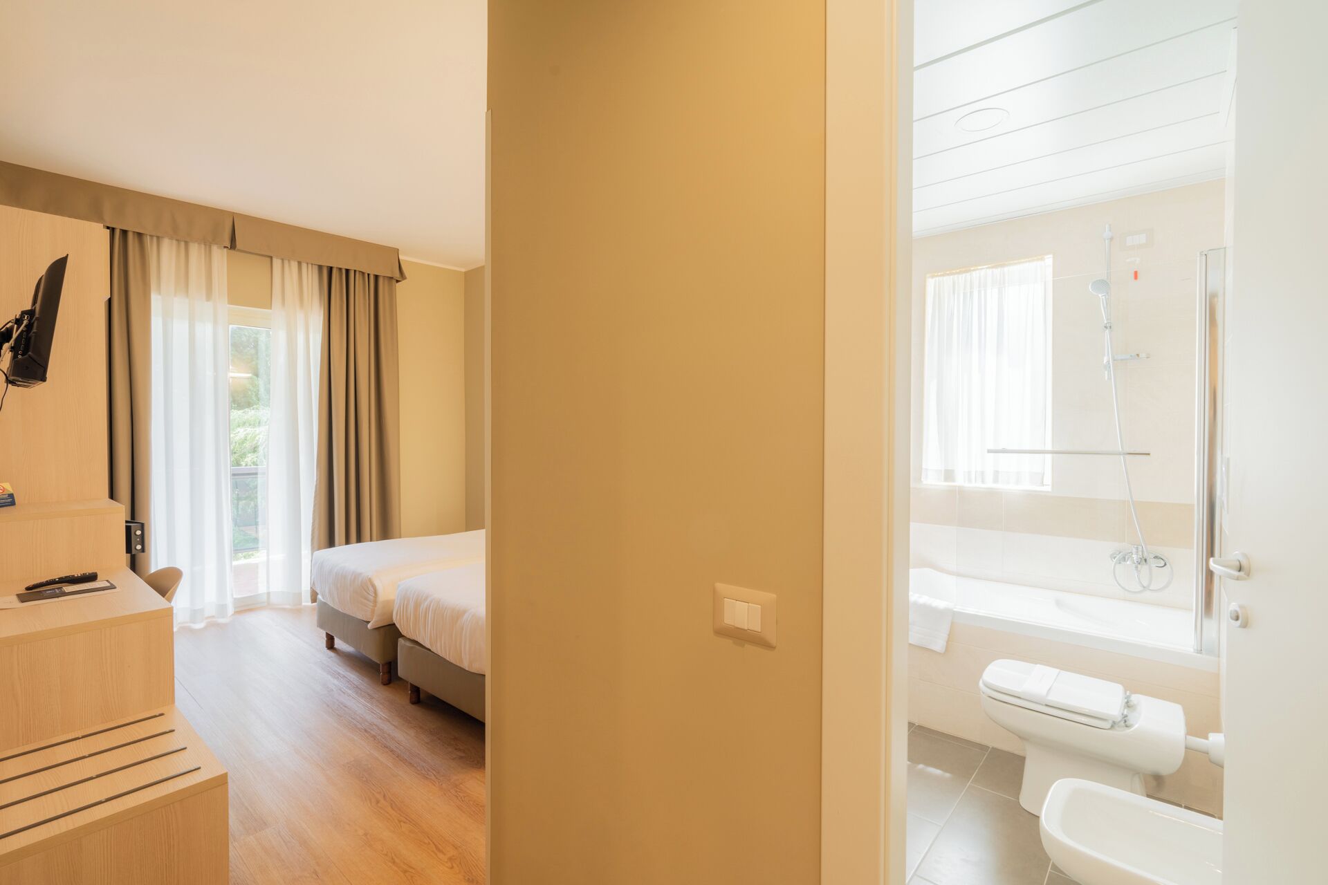 A hotel room with a bed, a desk with a computer, a bathroom with a shower, and a window with curtains.