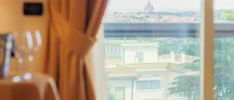 A hotel room with a bed, a window with curtains, and a view of buildings and greenery.