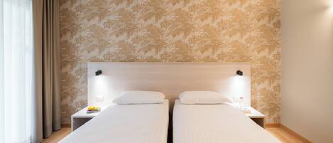 A hotel room with two single beds, a wooden headboard, a nightstand with a lamp, and a window with curtains.