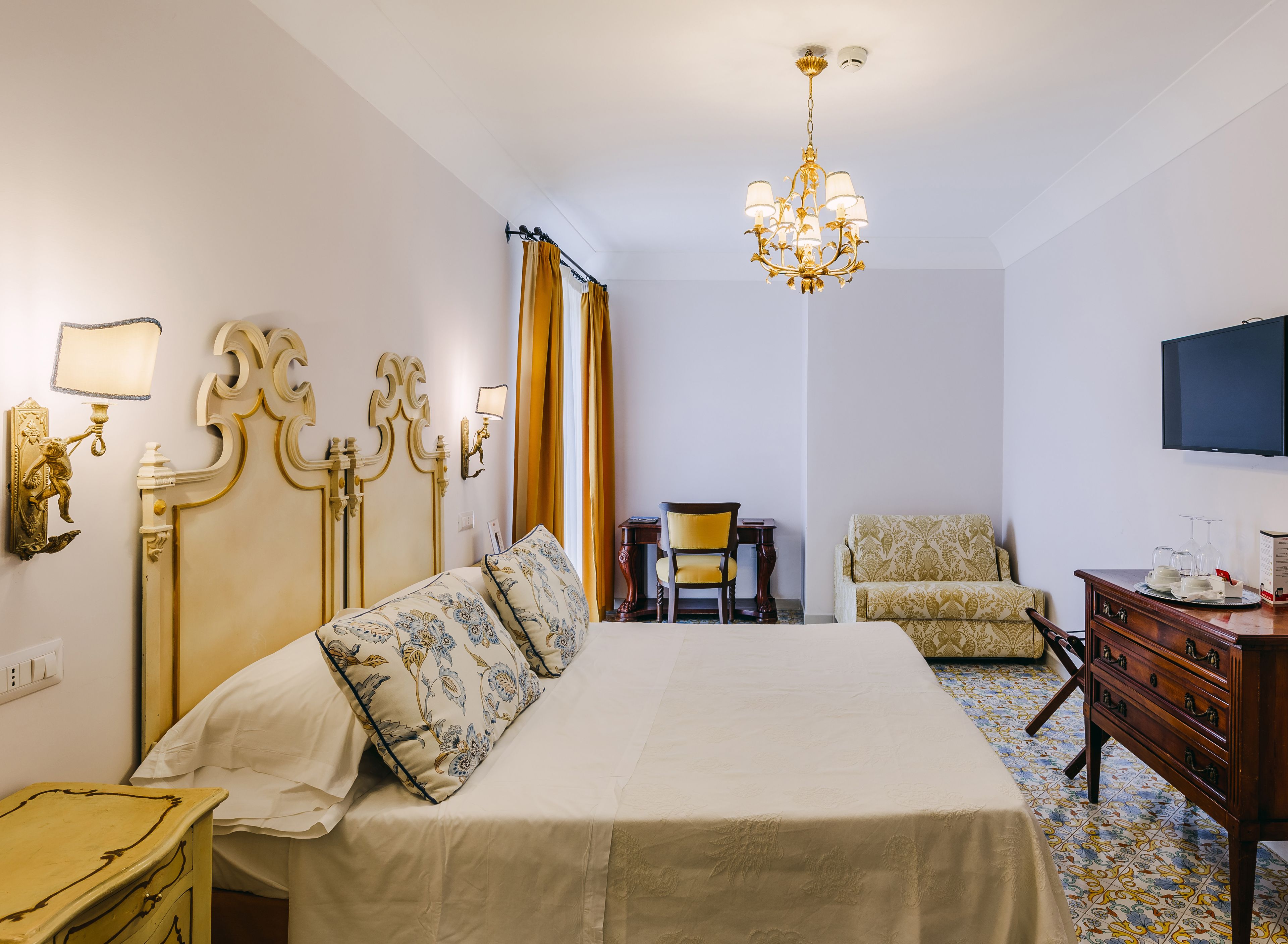 Une chambre d’hôtel avec un grand lit, un bureau, une chaise, une commode, un téléviseur et un lustre.