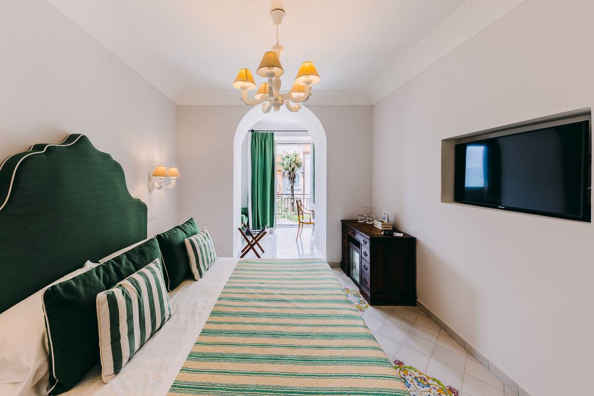 A hotel room with a large bed, a TV mounted on the wall, a chandelier, and a view of the outside balcony.