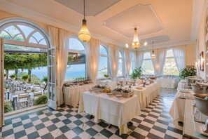 Un comedor con mesas dispuestas para una comida, ventanas amplias con cortinas y vista al mar.