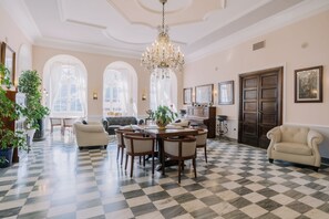Una habitación amplia con suelo a cuadros, un candelabro, una mesa de comedor con sillas, un sofá y un gran ventanal con arcos.
