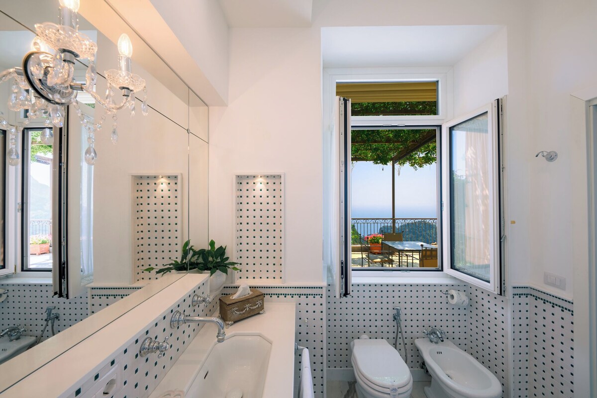 Un baño moderno con un espejo grande, una lámpara de araña, una bañera, un inodoro y un ventanal con vistas al exterior.