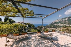 Una terraza con estructura metálica, plantas en macetas y vistas al mar y colinas.