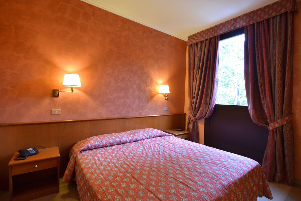 Habitación de hotel con cama, dos lámparas de noche, mesita de noche con teléfono y ventana con cortinas.