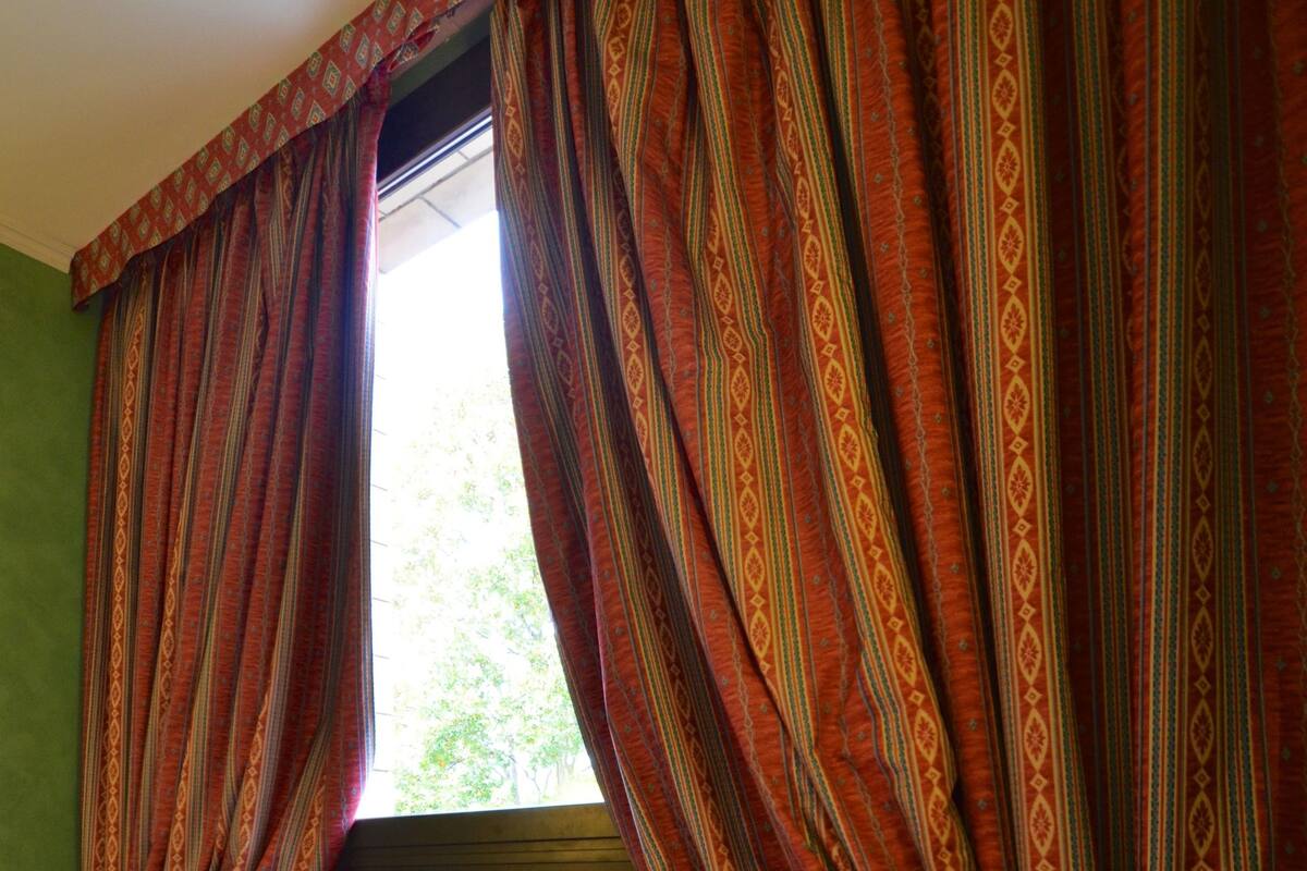 Una habitación con ventana cubierta por cortinas estampadas, suelo de madera y una sola silla.