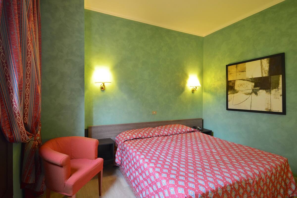 Una habitación de hotel con una cama, dos lámparas fijadas a la pared, un cuadro abstracto enmarcado y una cortina estampada.