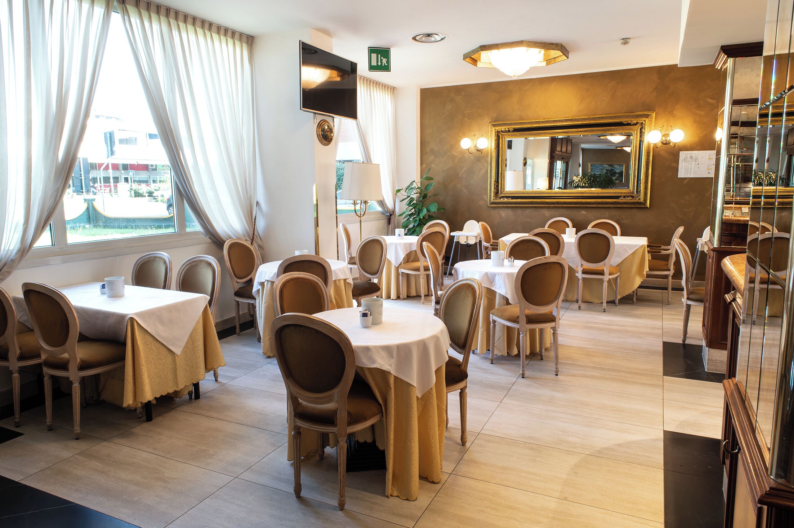 A hotel dining area with multiple tables and chairs, a large mirror, and a view of the outside through large windows.