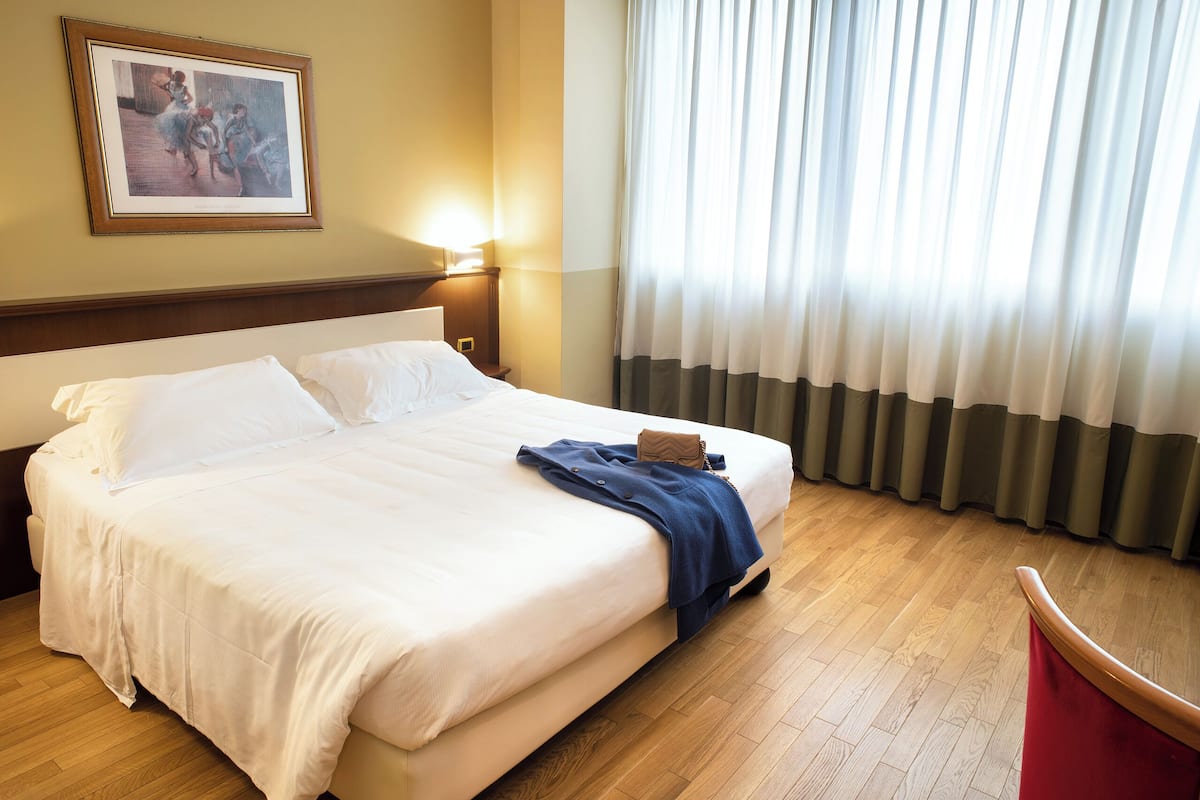 A hotel room with a large bed, a nightstand with a lamp, a framed picture on the wall, and a window with curtains.