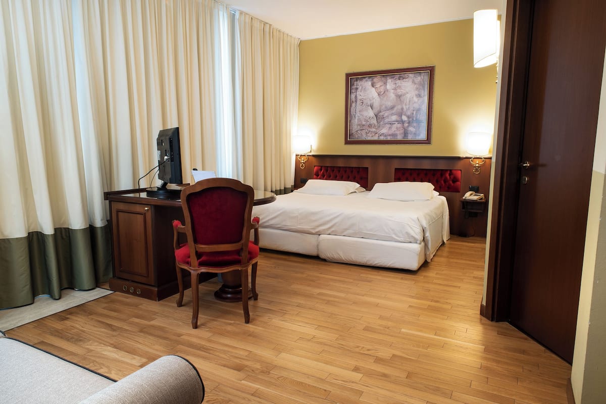 A hotel room with a wooden floor, a desk with a computer, a red chair, a bed with white bedding, and a painting on the wall.