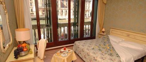 Habitación de hotel con cama, una mesa de comedor con fruta, una silla y vistas a edificios a través de un ventanal.