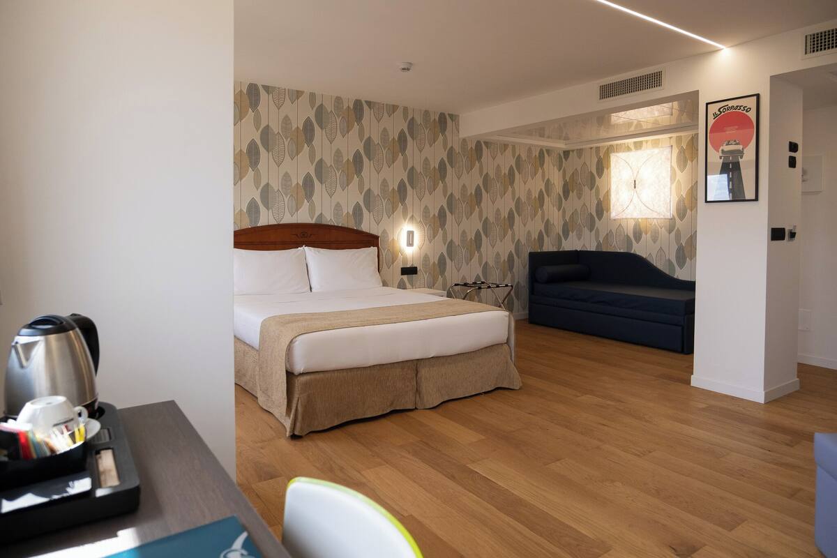 A hotel room with a bed, bedside table with a kettle and cups, a sofa, and a wall with a leaf-pattern wallpaper.